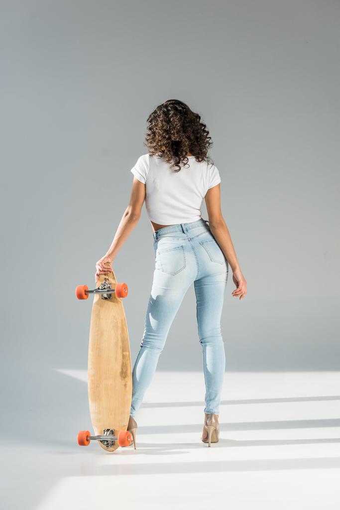 Achteraanzicht van vrouw met krullend haar reputatie met skateboard op grijze achtergrond - Foto, afbeelding