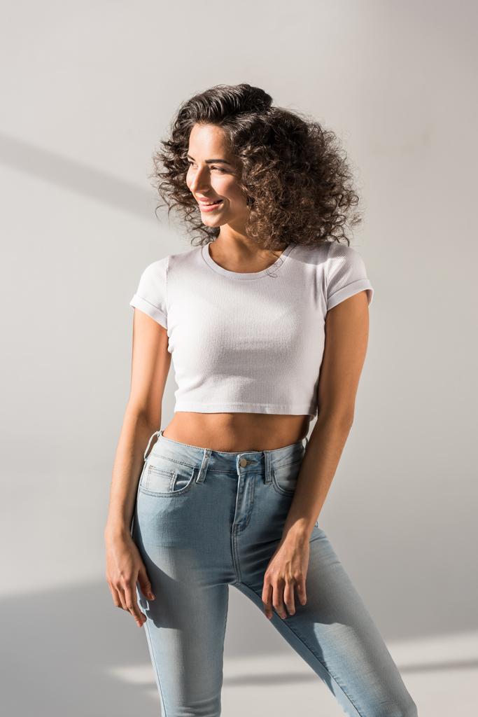 Smiling curly girl in jeans standing on grey background - Photo, Image
