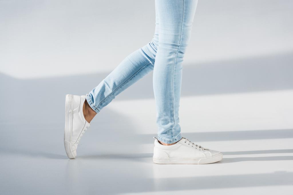 Gedeeltelijke weergave van vrouw in blauwe jeans en witte schoenen op grijze achtergrond - Foto, afbeelding