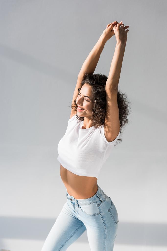 Excited curly woman in jeans posing with hands up on grey background - Photo, Image