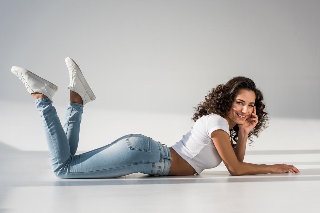 Pretty smiling girl in jeans lying on grey background - Photo, Image