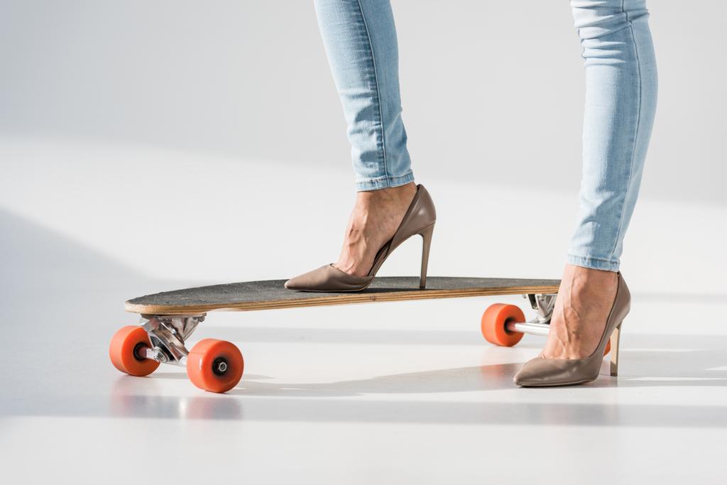 Vista cortada de mulher em jeans e sapatos de salto alto em pé sobre lonboard em fundo cinza
 - Foto, Imagem