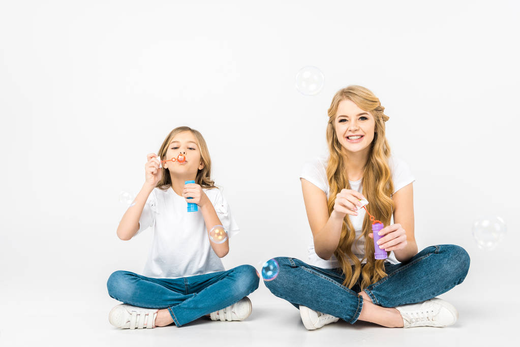 cheerful mother and daughter sitting on floor with crossed legs and blowing soap bubbles on white background - Photo, Image