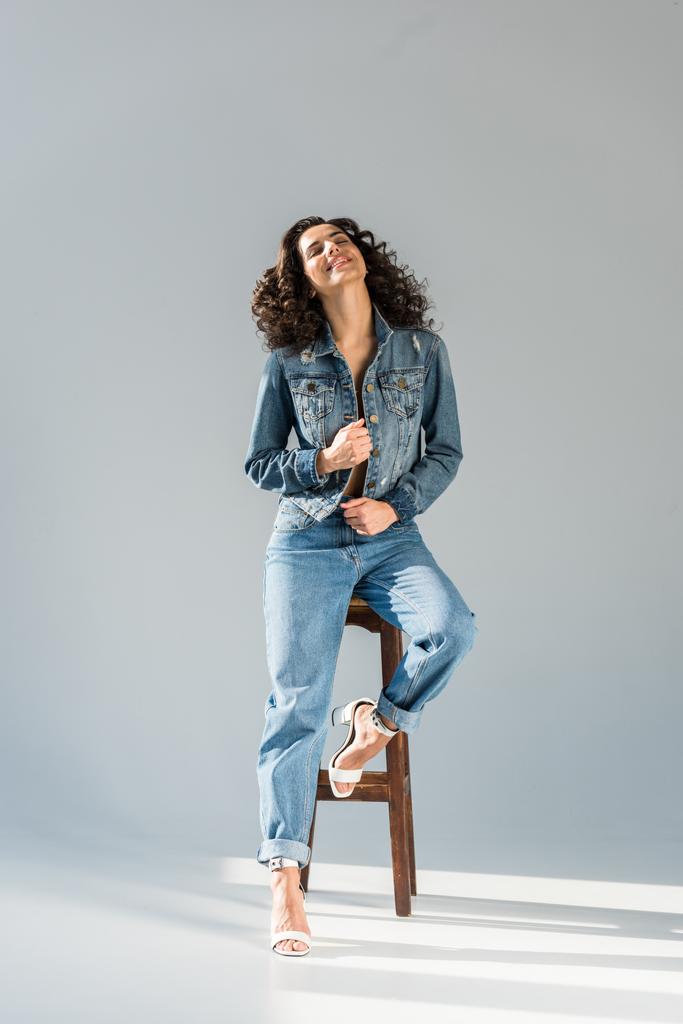 glückselige junge Frau in Jeans und Jacke sitzt auf einem Holzstuhl vor grauem Hintergrund - Foto, Bild