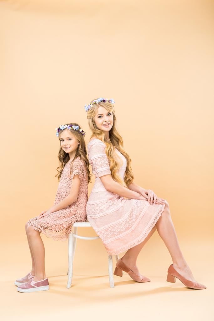 Szczęśliwa matka i córka w delikatne, koronkowe sukienki i kwiatowe wieńce siedzi na jednym krześle i patrząc na kamery na żółtym tle - Zdjęcie, obraz