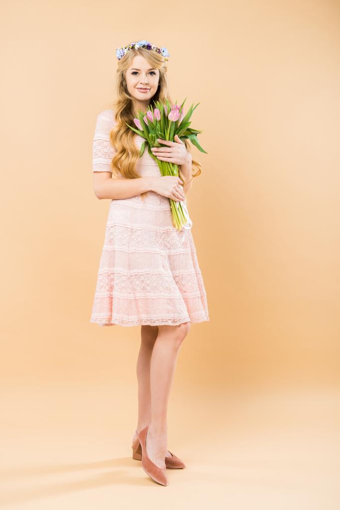 mooie vrouw in elegante kanten jurk en bloemen krans bedrijf boeket van roze tulpen op gele achtergrond - Foto, afbeelding