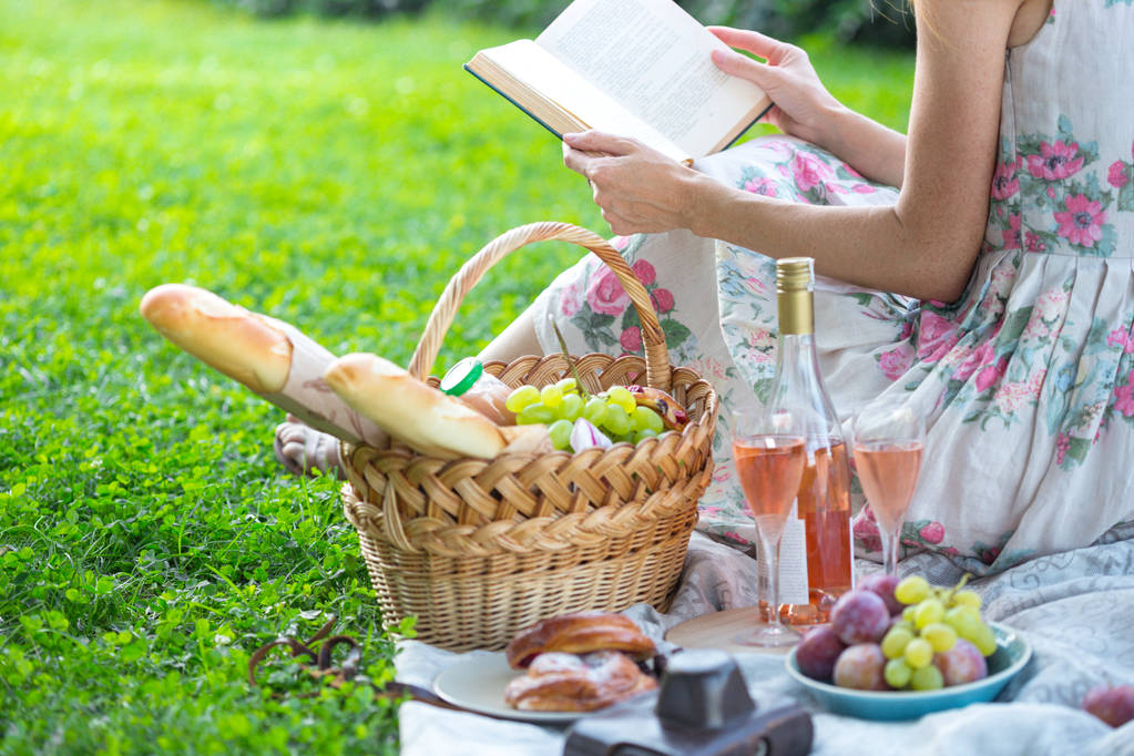 Lato - piknik na łące. dziewczyna siedzi czytając książkę i w pobliżu: piknik kosz i bagietki, wina, szklanki, winogron i rolki - Zdjęcie, obraz