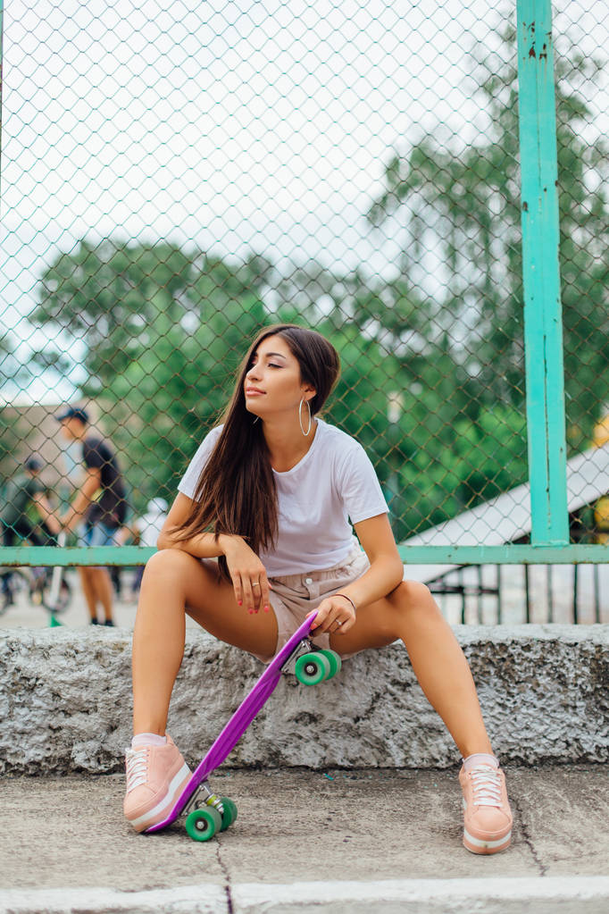 Sommer-Lifestyle-Bild von trendigen hübschen jungen Mädchen sitzt neben dem Skateboard-Wagen mit ihrem lila Plastik-Skateboard. - Foto, Bild