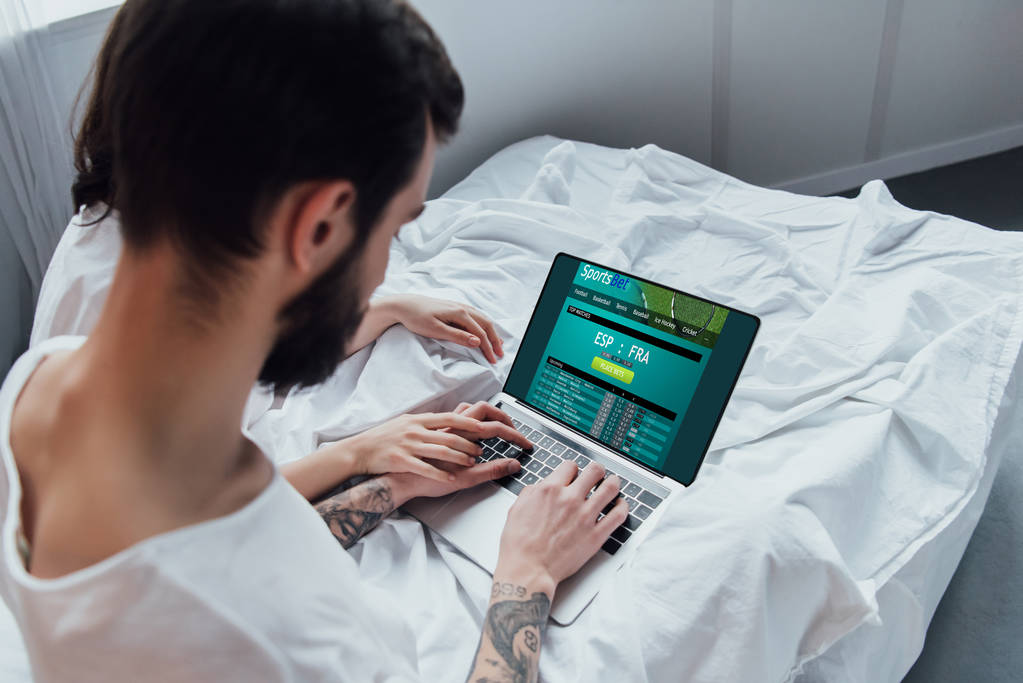 Rückansicht des Paares, das im Bett liegt, Hände hält und Laptop mit Sportwetten-Webseite auf dem Bildschirm benutzt  - Foto, Bild
