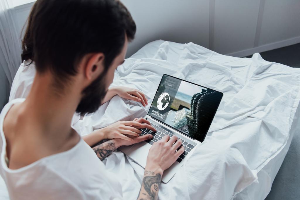 Achteraanzicht van paar liggend in bed, hand in hand en met behulp van laptop met boeking website op scherm  - Foto, afbeelding