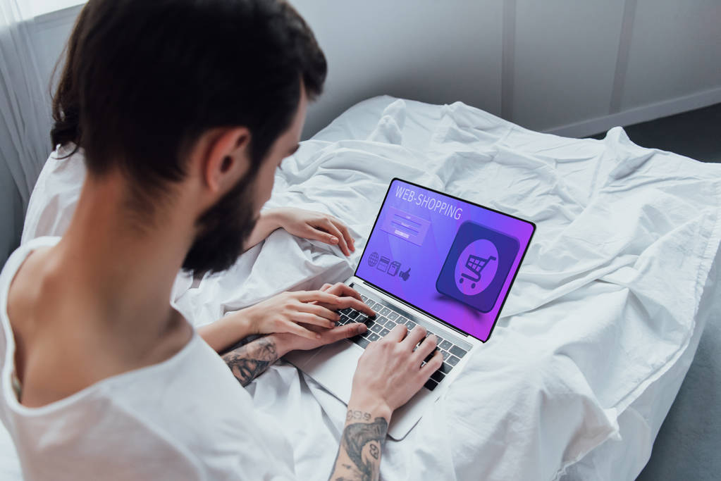 vista trasera de la pareja acostada en la cama, tomados de la mano y utilizando el ordenador portátil con el sitio web de compras en línea en la pantalla
  - Foto, imagen