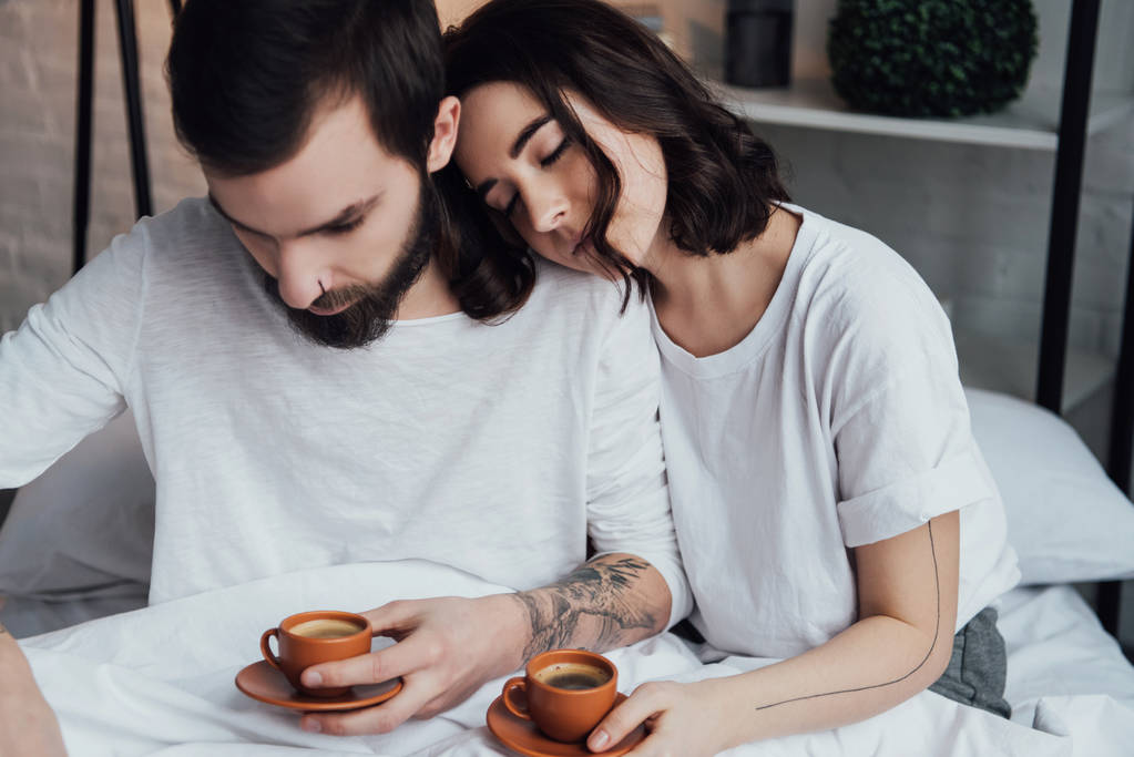 beautiful young tattooed couple lying in bed with coffee cups in morning - Photo, Image