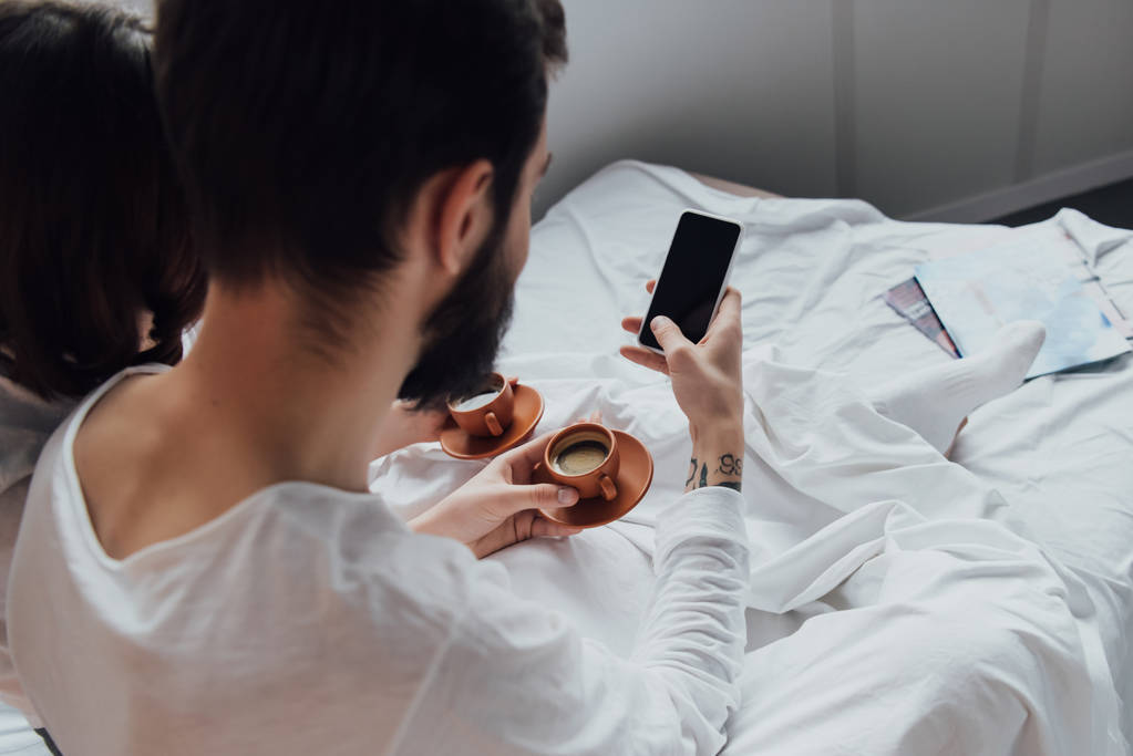 vista posteriore della giovane coppia sdraiata a letto con caffè e utilizzando smartphone con schermo bianco al mattino
 - Foto, immagini