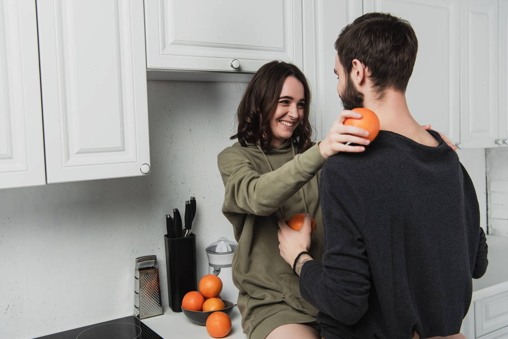 hermosa joven feliz pareja huyendo naranjas y mirándose en la cocina
 - Foto, imagen