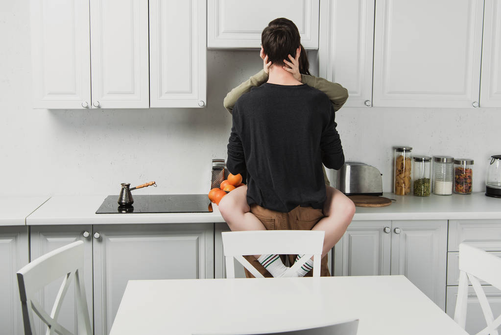 πίσω όψη του νεαρό ζευγάρι αγκάλιασμα και το φίλημα στο kithchen κατά τη διάρκεια του πρωινού - Φωτογραφία, εικόνα