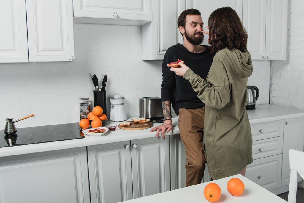 Frau hält Toast und wischt Mann beim Frühstück in Küche Marmelade aus dem Gesicht - Foto, Bild
