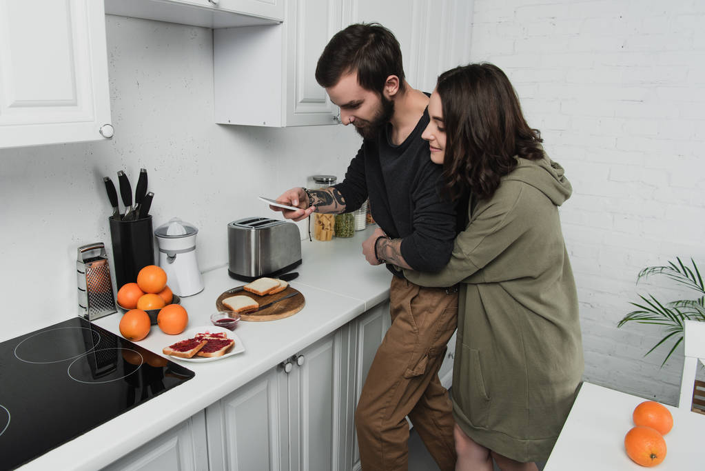 beautiful young couple taking photo of toasts on smartphone during breakfast in kitchen - Photo, Image