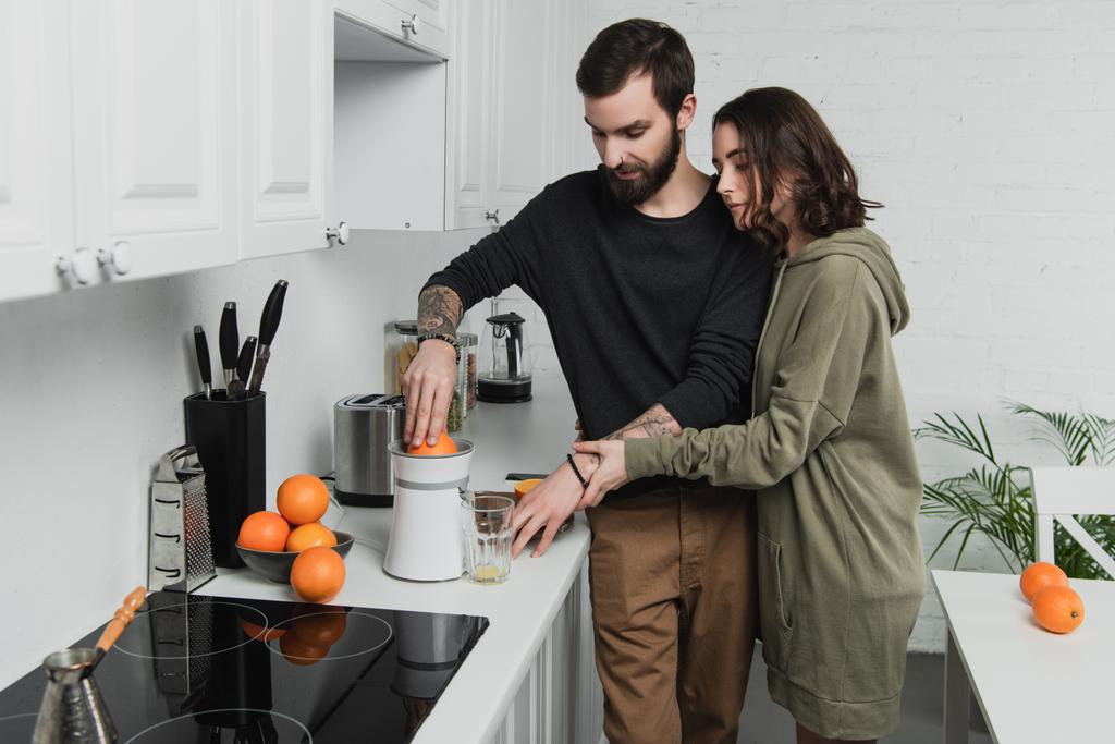 όμορφο νεαρό ζευγάρι προετοιμασία χυμό πορτοκαλιού κατά τη διάρκεια του πρωινού στην κουζίνα - Φωτογραφία, εικόνα