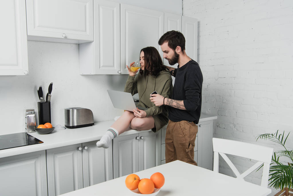 homme étreignant femme en utilisant un ordinateur portable et boire du jus d'orange pendant le petit déjeuner dans la cuisine
 - Photo, image