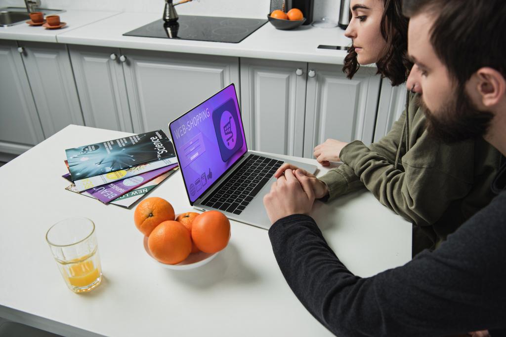 couple assis à table et à l'aide d'un ordinateur portable avec site de shopping en ligne à l'écran dans la cuisine
 - Photo, image