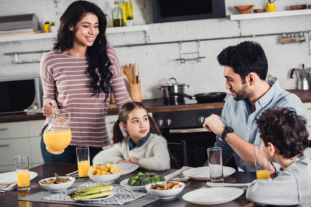fröhliche lateinische Mutter hält Glas mit Orangensaft in der Nähe von Tochter, Ehemann und Sohn während des Mittagessens - Foto, Bild