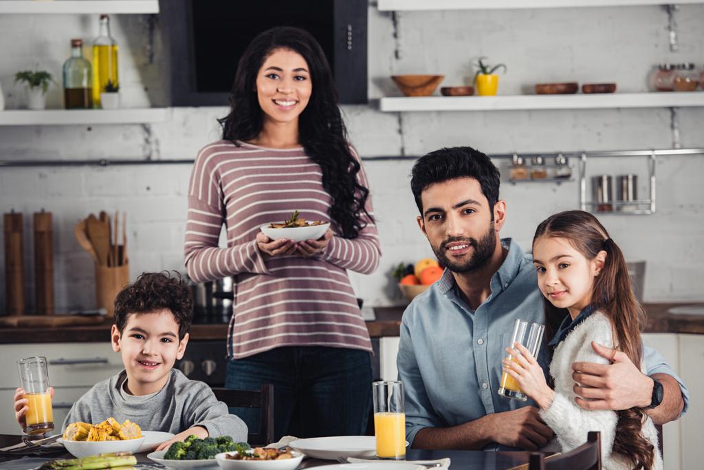 alegre madre latina sosteniendo plato con comida cerca de lindo hijo, marido e hija durante el almuerzo
 - Foto, imagen