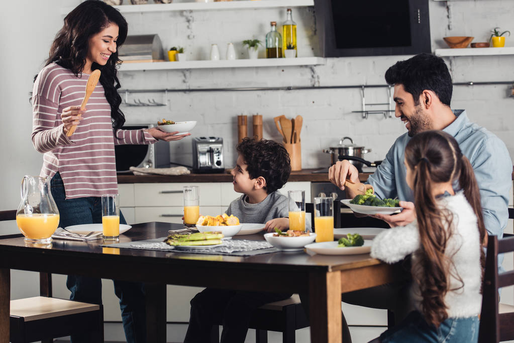 attractive latin woman looking at son while holding plate near daughter and husband during lunch - Photo, Image