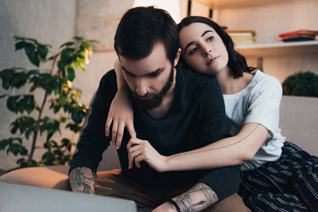 beautiful young tattooed couple embracing while sitting on couch in living room - Photo, Image