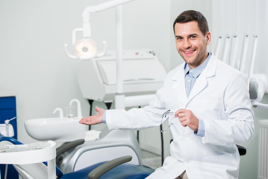 schöner Zahnarzt lächelt, während er in der Klinik gestikuliert - Foto, Bild