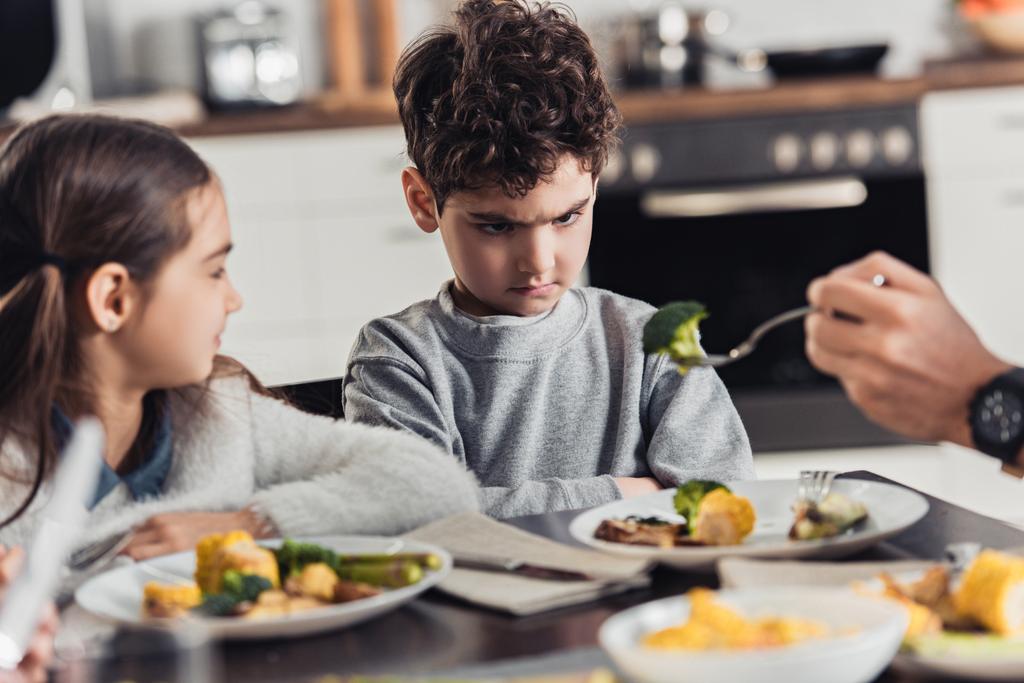 selectieve aandacht voor vork met groene broccoli in handen van vader met boos Latijns-jongen in de buurt van zus op achtergrond - Foto, afbeelding