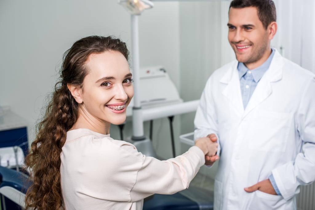 selective focus of cheerful woman in braces shaking hands with dentist standing with hand in pocket - Photo, Image