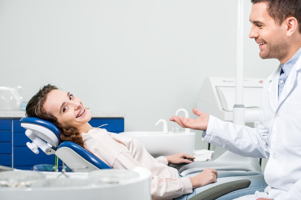 весела жінка в брекетах посміхається біля красивого стоматолога в клініці
 - Фото, зображення