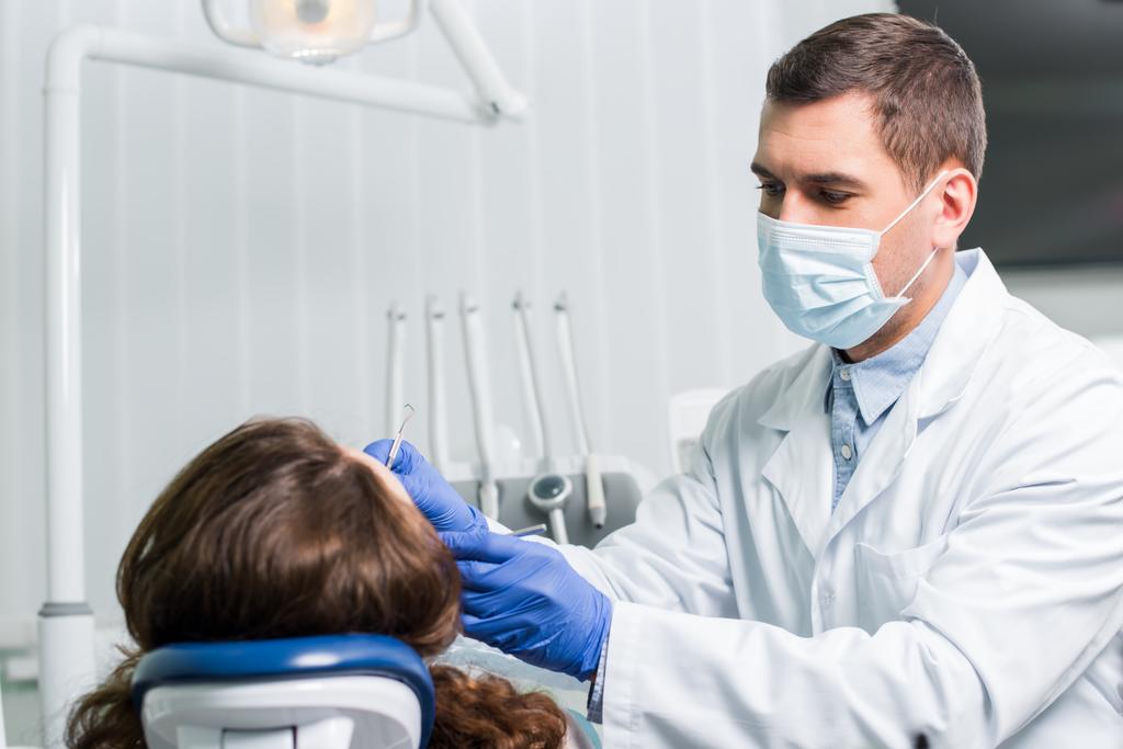 ernstige tandarts in latex handschoenen en een masker werken met vrouwelijke patiënt  - Foto, afbeelding