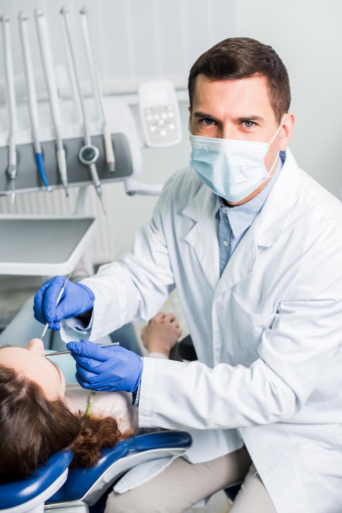 οδοντίατρος στο λάτεξ γάντια και μάσκα κρατώντας οδοντιατρικού εξοπλισμού κοντά θηλυκός ασθενής  - Φωτογραφία, εικόνα