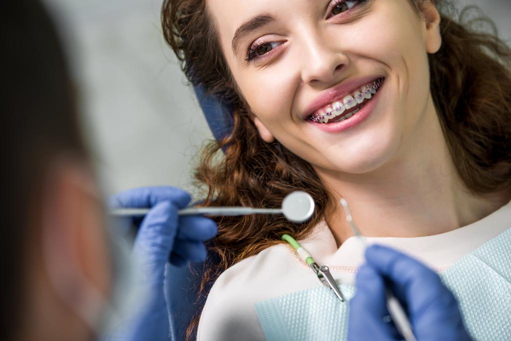 Close up van vrolijke vrouw tussen accolades tijdens onderzoek van tanden in de buurt van tandarts - Foto, afbeelding
