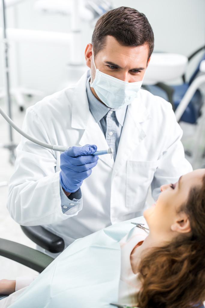 foco seletivo do dentista em jaleco branco e máscara segurando instrumento dentário perto de paciente do sexo feminino
 - Foto, Imagem