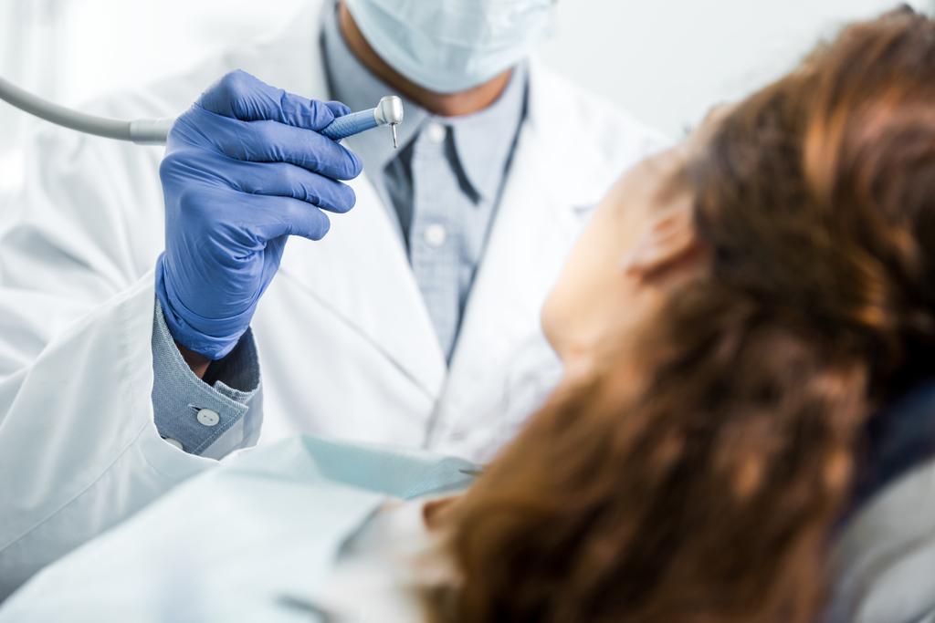περικοπεί θέα οδοντίατρο σε λευκό παλτό κρατώντας οδοντιατρικό όργανο κοντά θηλυκός ασθενής - Φωτογραφία, εικόνα