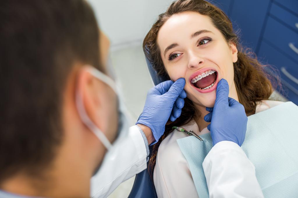 Selektywny fokus kobiety w nawiasy klamrowe, otwierając usta podczas badania zębów w pobliżu dentysta - Zdjęcie, obraz
