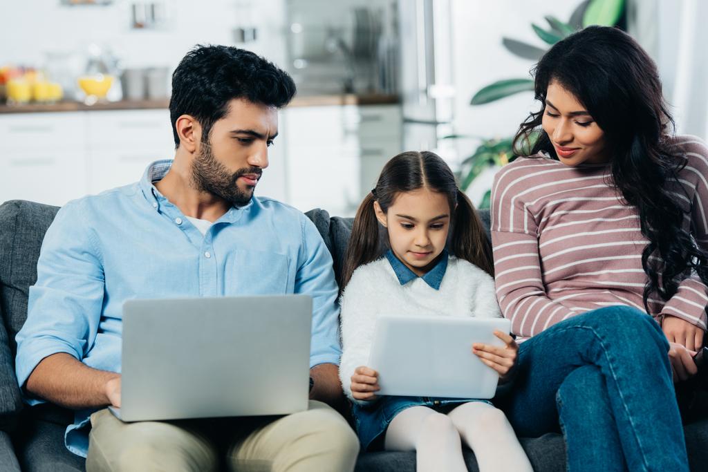handsome latin man with laptop looking at digital tablet in hands of cute daughter sitting near mother on sofa - Photo, Image