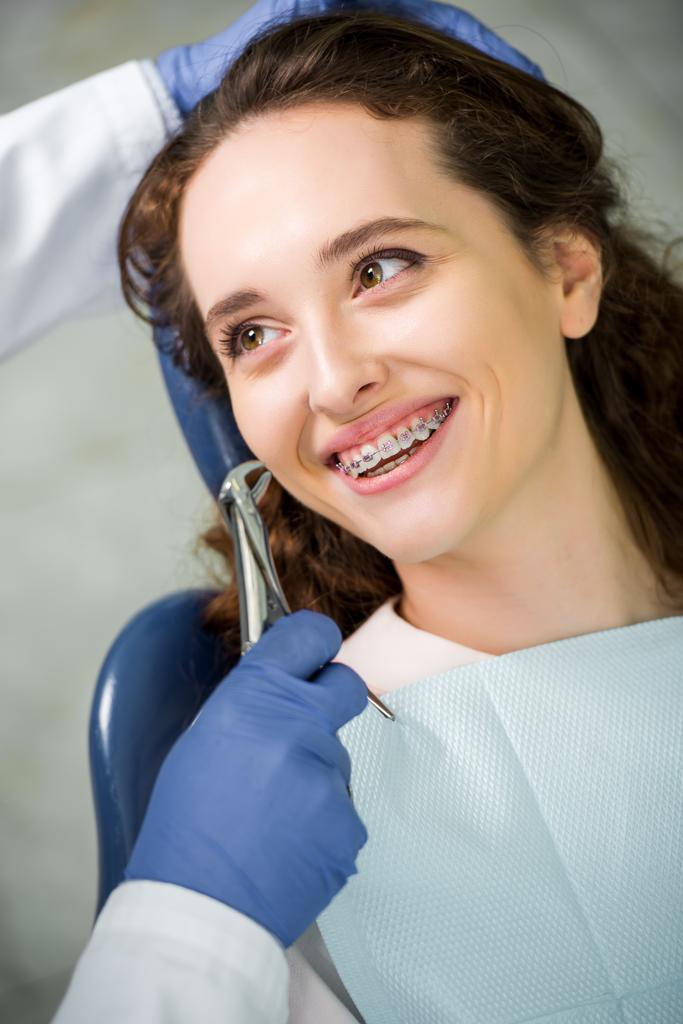 Selektywny fokus kobiety w nawiasy klamrowe, uśmiechnięty patrząc na stomatologa podczas badania  - Zdjęcie, obraz