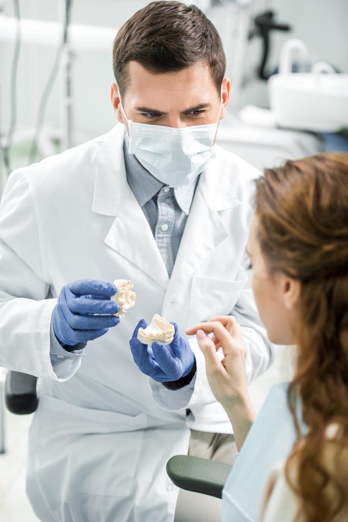 Selektiver Fokus des Zahnarztes in Latexhandschuhen und Maske, die das Zahnmodell in der Nähe der Patientin hält - Foto, Bild