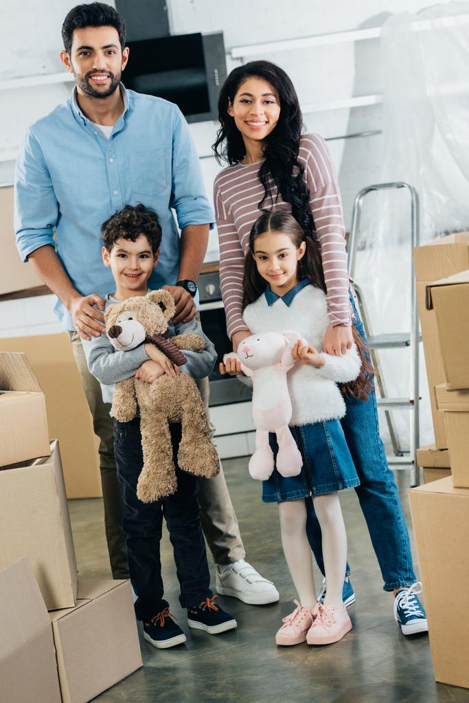 happy latin kids holding soft toys and standing with cheerful mom and dad in new home - Photo, Image