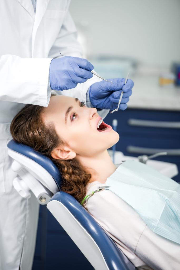 oříznutý pohled zubař v latexové rukavice zkoumání žena v závorkách s otevřenou hubou  - Fotografie, Obrázek