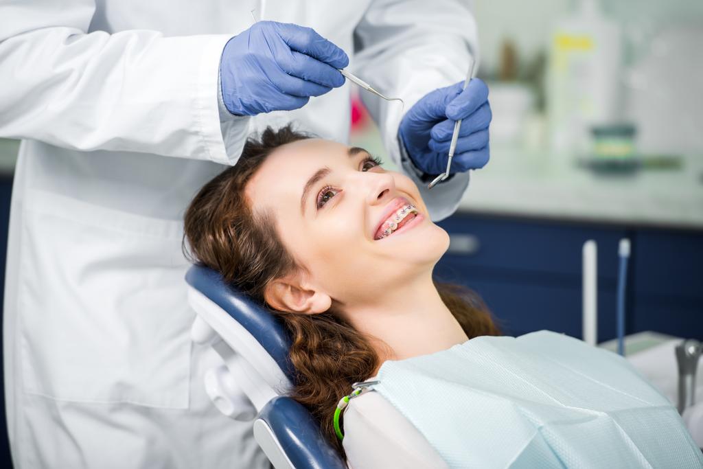 Ausgeschnittene Ansicht eines Zahnarztes in Latexhandschuhen, der eine fröhliche Frau in Zahnspange untersucht  - Foto, Bild