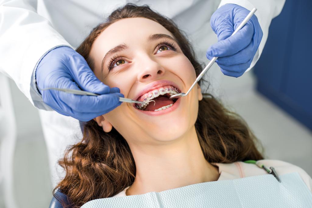 vista ritagliata del dentista in guanti di lattice esaminando donna allegra in bretelle con bocca aperta
  - Foto, immagini