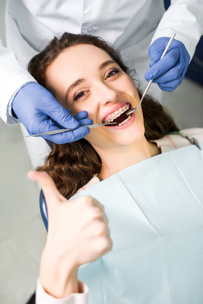 Ausgeschnittene Ansicht des Zahnarztes in Latexhandschuhen bei der Untersuchung einer fröhlichen Frau in Zahnspange mit geöffnetem Mund, die den Daumen nach oben zeigt  - Foto, Bild