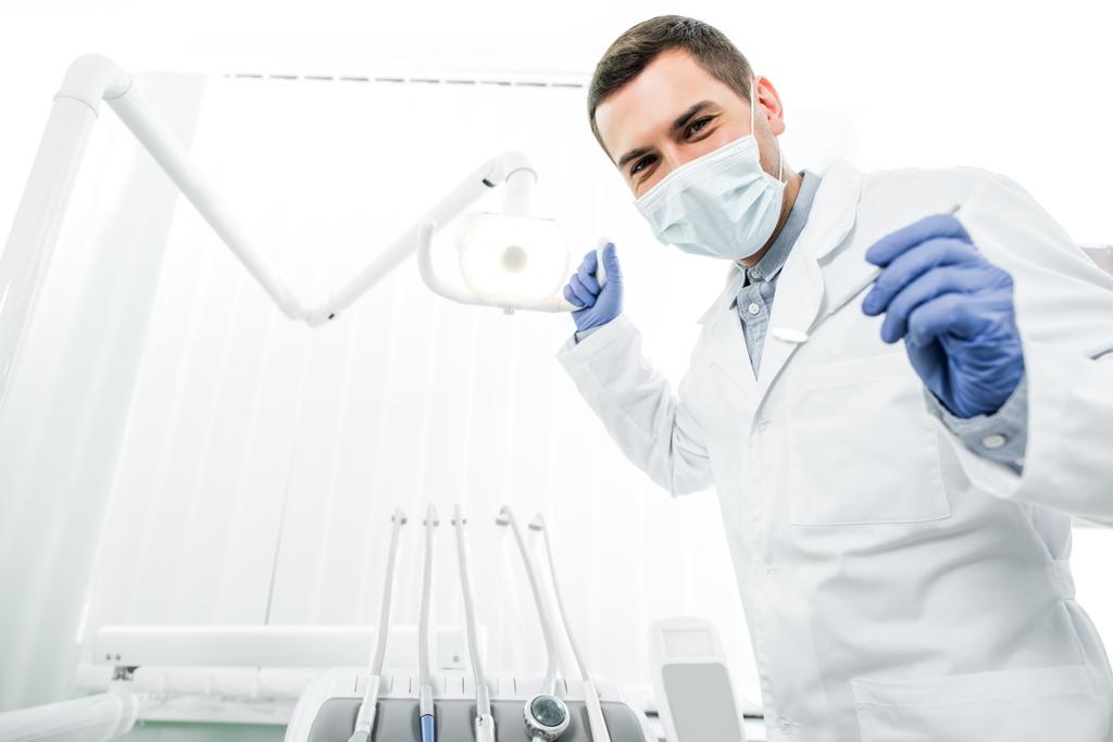 dentista en guantes de látex y máscara que sostiene el instrumento dental y la lámpara dental
 - Foto, imagen