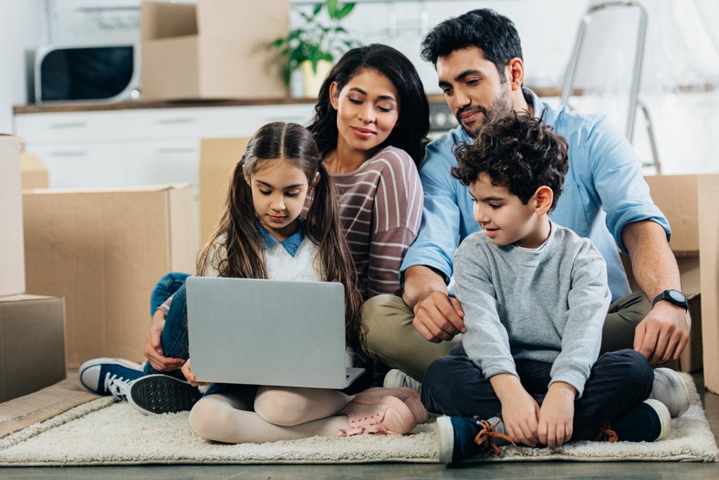 χαρούμενα latino οικογένεια ψάχνει στο φορητό υπολογιστή ενώ κάθεται στο χαλί στο νέο σπίτι - Φωτογραφία, εικόνα