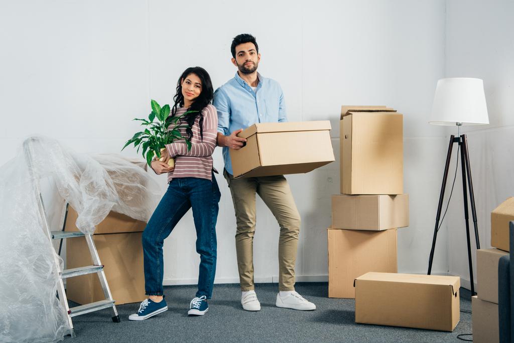 счастливая латинская женщина держит растение рядом с мужем с коробкой во время переезда в новый дом
 - Фото, изображение