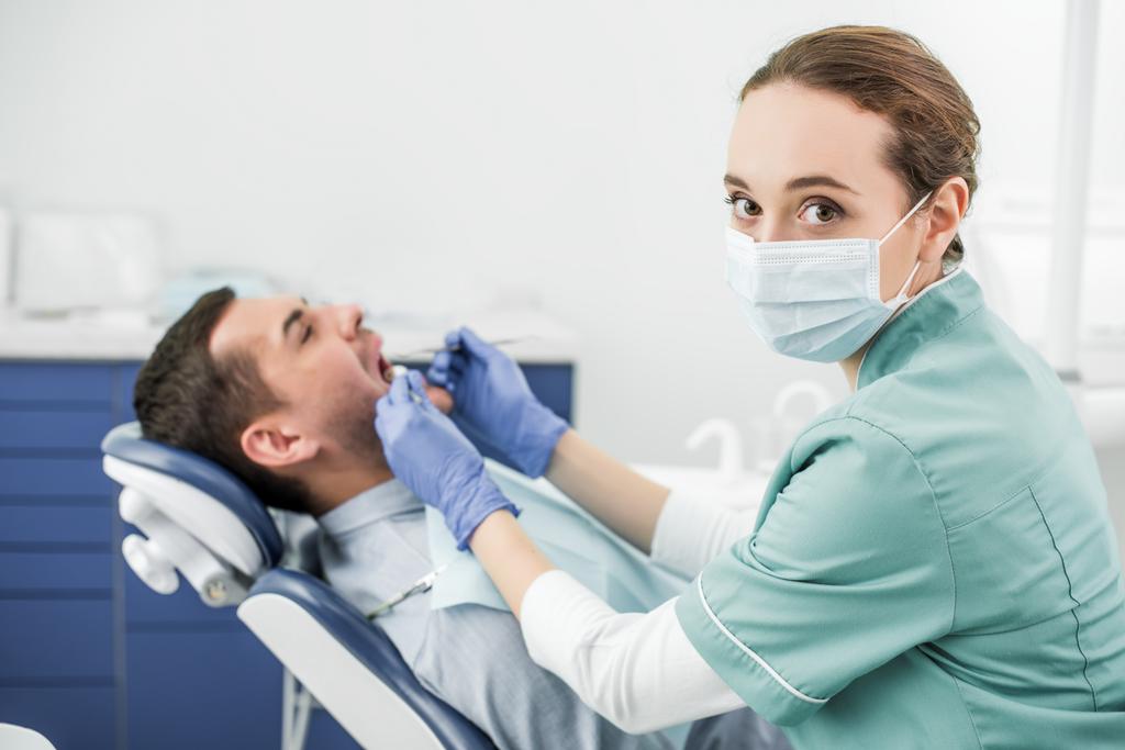 επιλεκτική εστίαση του οδοντιάτρου βλέπουν φωτογραφική μηχανή και κρατώντας οδοντικά όργανα κοντά σε ασθενή με ανοικτό στόμα - Φωτογραφία, εικόνα